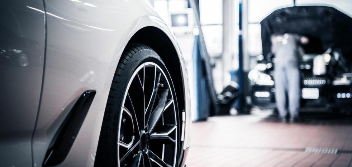 Close up of a car alloy wheel while a man works on the engine of another car in the background