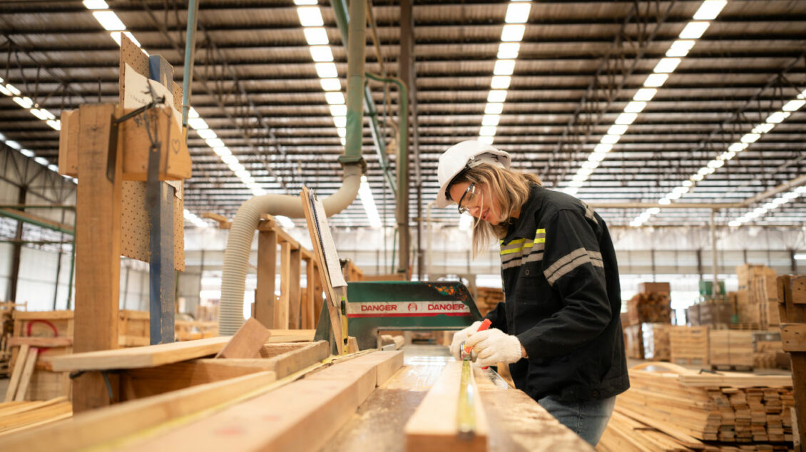 A woman in a hard hat measuring and cutting timber in a factory